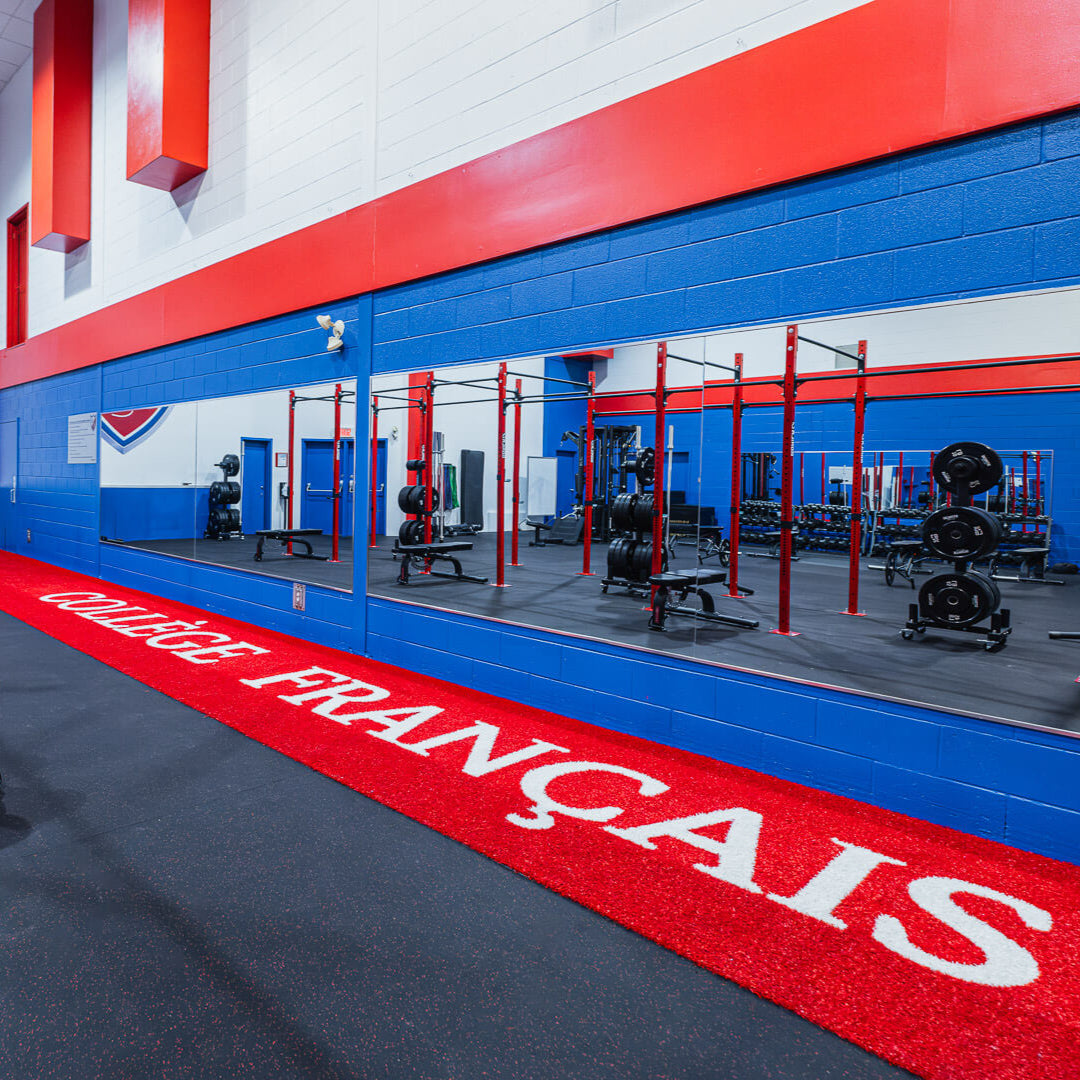 A sleek gym facility with vibrant red and blue accents. The mirrored wall reflects a functional training area featuring red and black workout rigs, racks, and weightlifting equipment. The bold, custom red turf proudly displays the name "Collège Français" in large white letters, adding a professional and branded touch to the space.