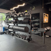 A black and gray multi function gym equipment storage rack filled with kettlebells, bumper plates and medicine balls surrounded by exercise equipment in a dark gym interior.