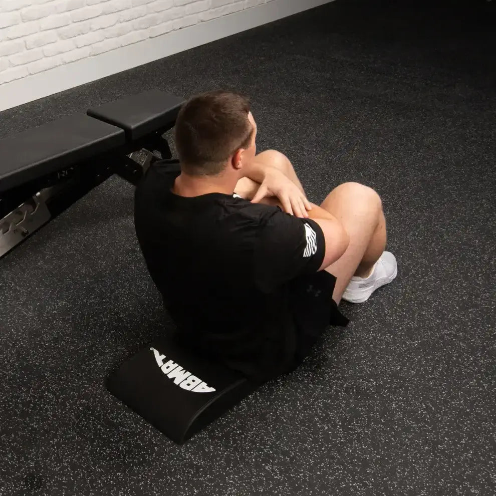 Person using a black exercise abmat on a gym floor with a white brick wall in the background.