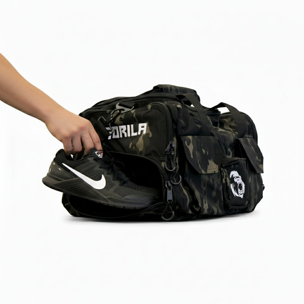 Athlete putting his Nike Metcons shoes in the shoe compartment of the Gorila Gym Bag in camouflage pattern, side view with visible logo and accessory pockets.