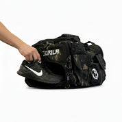 Athlete putting his Nike Metcons shoes in the shoe compartment of the Gorila Gym Bag in camouflage pattern, side view with visible logo and accessory pockets.